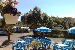 Campingplatz mit Stellplätzen, Wohnwagen und blauen Zelten, Sitzgruppe und Bäumen im Hintergrund.