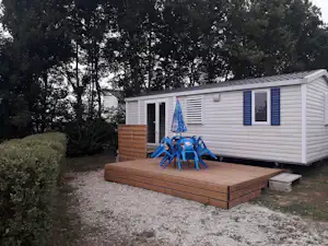 Mobilheim mit Holzterrasse, blauen Markisen und Stühlen vor einem Weg, umgeben von Bäumen im Campingbereich.