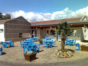 Terrasse am Campingplatz mit blauen Kunststoffstühlen und Tischen, Steinpflaster, Pflanzenkübeln und Gebäudefassade unter Vordach.