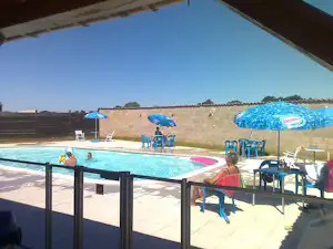 Camping-Pool mit blauem Sonnenschirm, Liegen und Tischen auf der Terrasse, im Hintergrund eine Steinmauer.
