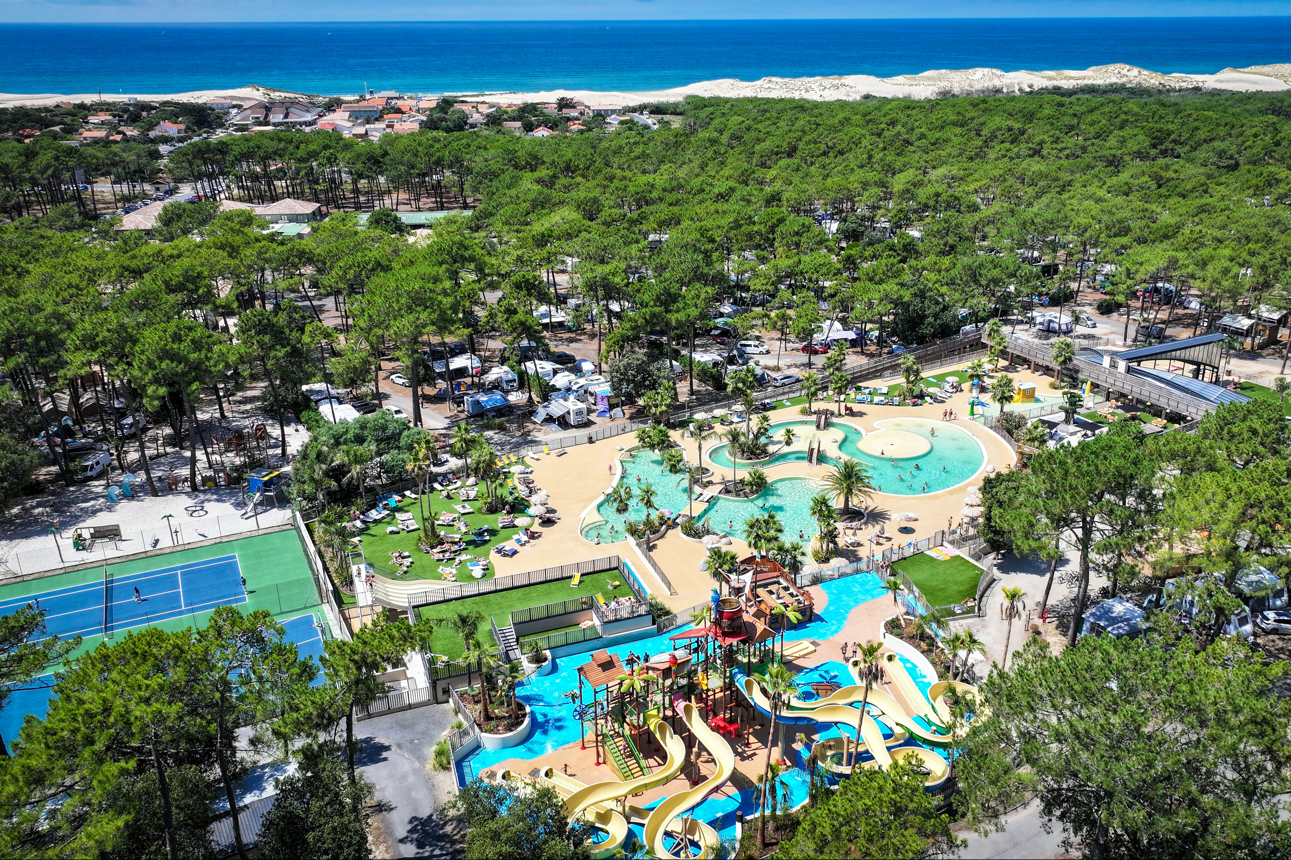 Großes Campinggelände mit mehreren Pools, Rutschen und Liegen, umgeben von Bäumen und kleinen Wegen, nahe am Meer.