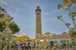Leuchtturm mit roter Laterne, im Vordergrund Blumen und Dächer eines Cafés, dahinter Himmel mit Wolken.