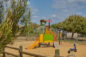 Bunter Spielplatz mit Kletterturm und Rutsche, Sandboden und Holzgeländer, umgeben von Bäumen und Sträuchern.