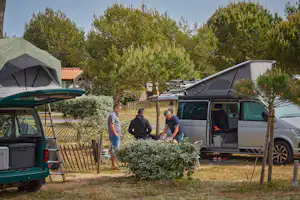 Wohnmobil und Campingbus am Stellplatz, daneben Zelte und ein Zaun; im Hintergrund Bäume und ein kleiner Weg.