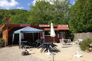 Terrasse am Camping mit Snack-Stand, Schirm und Sitzgruppe vor Schildern für Pizzas, Snack und Frites.