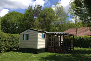 Mobilheim mit Veranda und Holzgeländer im Grünen, umgeben von Bäumen und Rasenfläche.