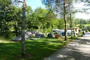 Zelte und Wohnwagen auf einem Campingplatz im Grünen, umgeben von Bäumen und einem Kiesweg.