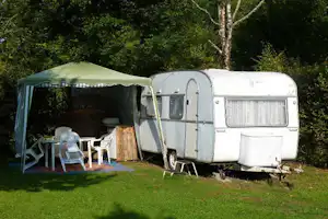 Wohnwagen mit Vorzelt auf einem Campingplatz, daneben weiße Stühle und Tisch auf dem Stellplatz im Grünen.