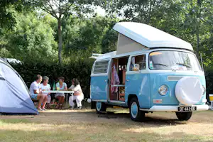 Blaues Wohnmobil mit geöffnetem Fenster und Dachhaube, daneben Zelt und Tischgruppe auf einem Campingplatz im Grünen.