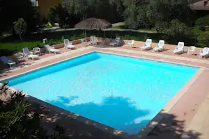 Camping-Pool mit blauem Wasser, Terrassenboden und mehreren weißen Liegestühlen, im Hintergrund Sonnenschirm und Hecke.