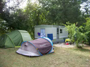 Zelte auf einem Campingplatz neben einem Mobilheim, mit Grasfläche, Bäumen und einem kleinen Tisch am Stellplatz.