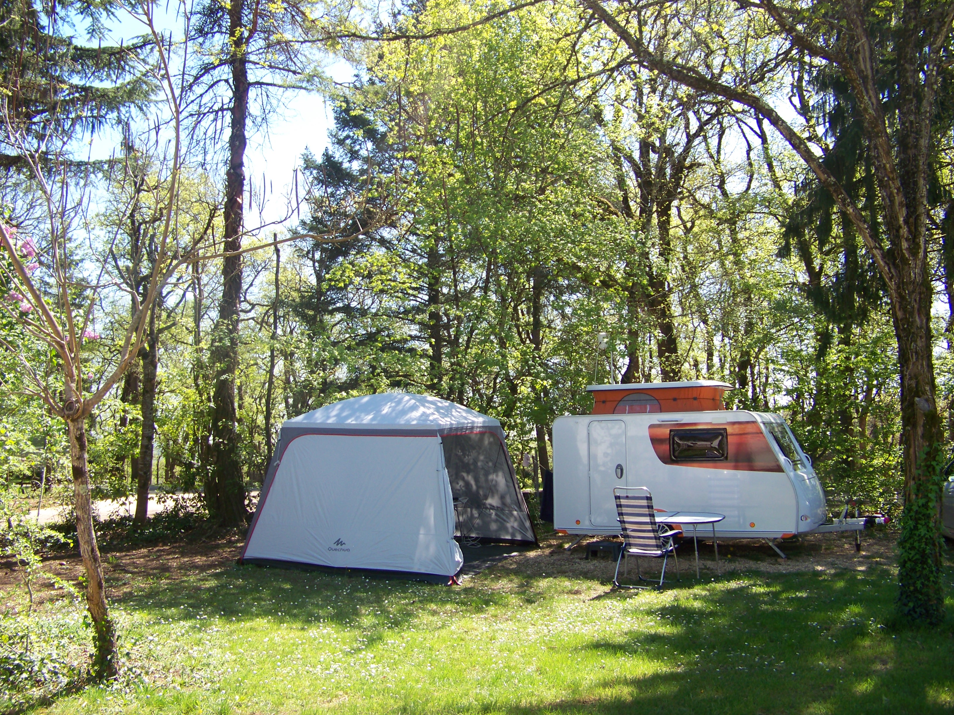 Campingzelt und Wohnwagen auf einer grünen Wiese im Schatten von Bäumen, mit Stühlen und Tisch am Wohnwagen.