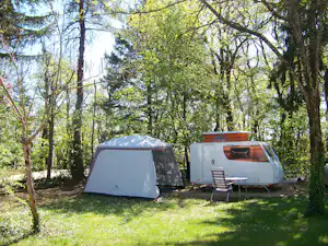 Campingzelt und Wohnwagen auf einer grünen Wiese im Schatten von Bäumen, mit Stühlen und Tisch am Wohnwagen.