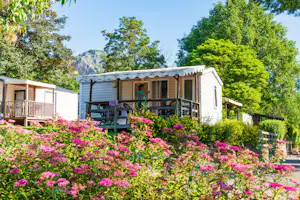 Mobilheim mit Terrasse und Holzgeländer, davor blühende pinke Blumenbeete und Bäume, im Hintergrund Felswand.