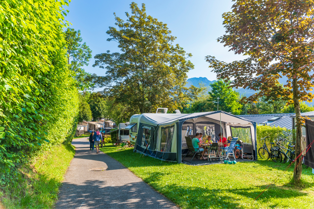 Großes Zelt mit Sitzgruppe und Fahrrädern am Campingplatz, daneben Wohnwagen und ein Weg zwischen Bäumen.