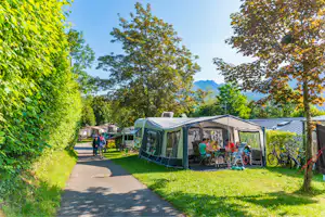 Großes Zelt mit Sitzgruppe und Fahrrädern am Campingplatz, daneben Wohnwagen und ein Weg zwischen Bäumen.