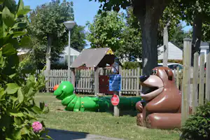 Spielplatz auf dem Campingplatz mit grüner und brauner Tierfigur, Holzzaun, Bäumen und Rasen im Hintergrund.