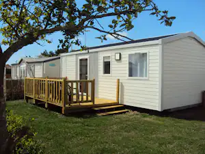 Mobilheim mit Holzveranda und Eingangstür, Rasenfläche davor, Hecke und Bäume im Hintergrund.