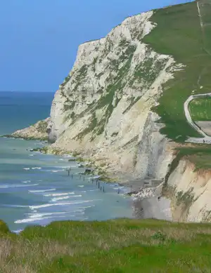 Kreidefelsen an der Küste mit Sandstrand, Wellen und einem Küstenweg am Hang.