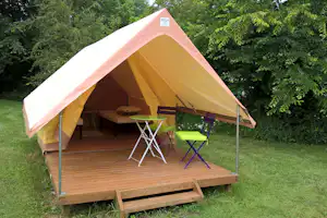 Gelbes Zelt auf Holzterrasse mit Tisch und grünen Stühlen, umgeben von Bäumen und dichtem Grün.