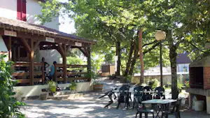 Terrasse am Campingplatz mit Tischen und Stühlen vor einer überdachten Holzbar „Snack-bar“ unter Bäumen.