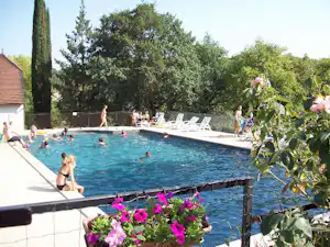 Camping-Pool mit blauem Wasser, Liegen und Blumen im Vordergrund, Bäume und Gebäude am Rand.