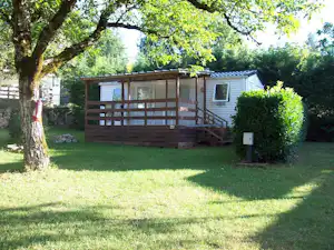 Mobilheim mit Holzveranda und Treppe im grünen Campinggarten, umgeben von Bäumen und Hecken.