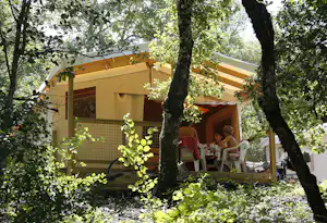 Mobilheim im Grünen mit Terrasse, Stühlen und Tisch, umgeben von Bäumen im Campingbereich.