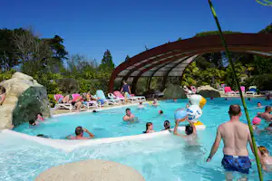 Großes Camping-Schwimmbecken mit Wasserrutsche, Liegen und mehreren Personen im Wasser, im Hintergrund Bäume und Gebäude.