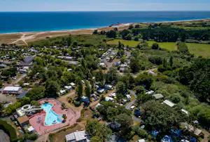 Luftaufnahme eines Campingplatzes mit Pool, Wohnwagen und Bungalows, umgeben von Bäumen, nahe dem Meer und einem Strandstreifen.