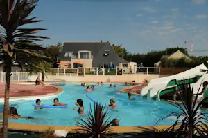 Großer Camping-Pool mit Wasserrutsche, Menschen im Becken und weißen Wohngebäuden im Hintergrund, Palmen und Liegen am Rand.