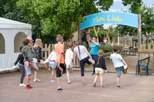 Kinder laufen unter dem Miniclub-Schild auf dem Campingplatz, im Hintergrund Zelte, Bäume und ein Holzzaun.