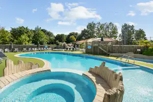 Schwimmbecken mit Whirlpool-Ecke, Liegen und Sonnenschirmen am Campingplatz, umgeben von Bäumen und Holzsichtschutz.