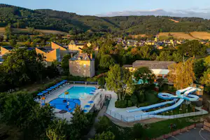 Campinganlage mit blauem Schwimmbecken, Liegen und Sonnenterrasse, umgeben von Bäumen und Häusern in einer hügeligen Landschaft.