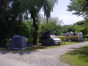 Zelt und Wohnmobil auf einem Campingplatz, umgeben von Bäumen, mit Kiesweg und Rasenfläche im Hintergrund.