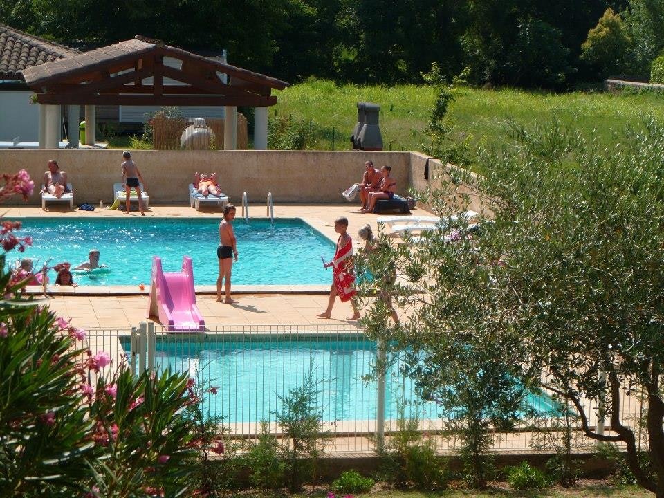 Schwimmbad auf dem Campingplatz mit mehreren Liegen, Sonnenschirmen und einer Terrasse am Pool, umgeben von Bäumen und Grün.