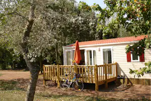 Mobilheim mit Holzterrasse, orangefarbenem Sonnenschirm und Fahrrad vor weißen Wänden, umgeben von Bäumen und Gartenweg.