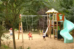 Spielplatz mit Klettergerüst, Rutsche und Schaukeln im Sand, umgeben von Bäumen und grünem Buschwerk.