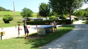 Campingplatz mit Wohnwagen, Grilltisch und Personen am Wegesrand, Bäume und gepflegte Rasenflächen im Hintergrund.