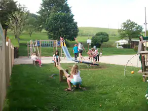 Spielplatz auf dem Campingplatz: Kinder und Erwachsene am Rutschenturm, daneben Rasen, Bäume und ein Wohnwagen in der Ferne.