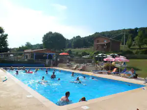 Großer Camping-Pool mit Liegen, Sonnenschirmen und mehreren Personen im Wasser, im Hintergrund ein Gebäude und grünes Gelände.