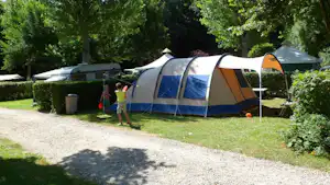 Campingzelt auf einem Grasplatz, daneben Wohnwagen und weitere Zelte, Hecken und Bäume im Hintergrund.