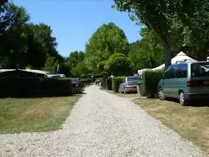 Schotterweg auf einem Campingplatz zwischen Hecken und parkenden Autos, mit Bäumen und Rasenflächen am Rand.