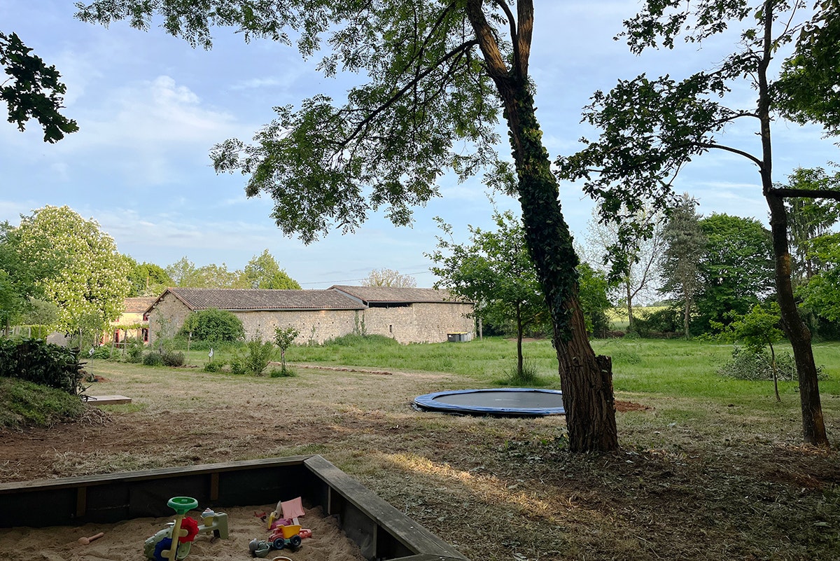 Campingplatz mit Rasenfläche, Bäumen und einem kleinen Spielbereich am Rand, im Hintergrund Gebäude und Wege.