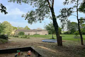 Campingplatz mit Rasenfläche, Bäumen und einem kleinen Spielbereich am Rand, im Hintergrund Gebäude und Wege.