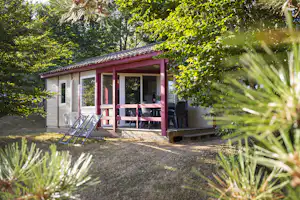 Bungalow mit Holzterrasse und Sitzgruppe vor dem Eingang, umgeben von Bäumen und Sträuchern im Grünen.