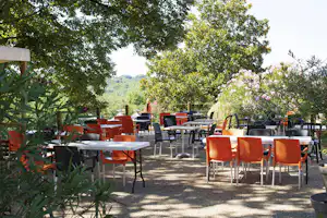 Terrasse mit Tischen und orangefarbenen Stühlen, umgeben von Bäumen und Sträuchern, mit Blick auf grüne Hügel.