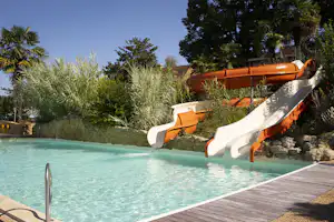 Pool mit orangefarbener Rutsche am Campingplatz, Holzsteg und Palmen im Hintergrund.