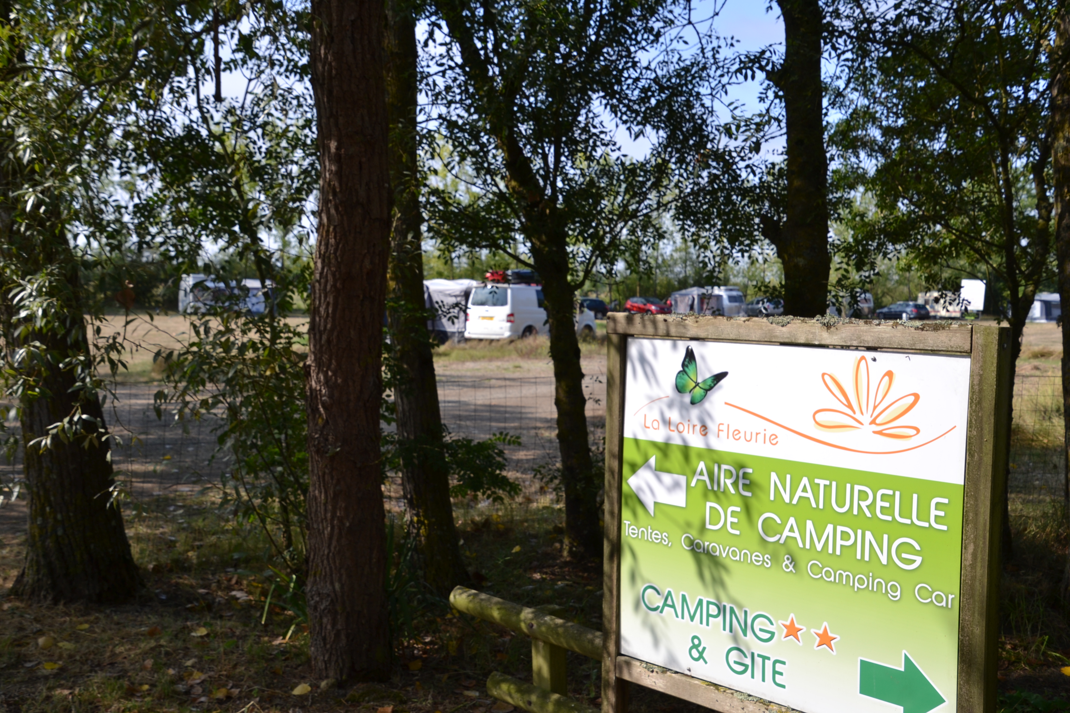 Camping-Schild „Aire Naturelle de Camping“ mit Text und Logo, im Hintergrund Wohnwagen und Bäume im Campingbereich.