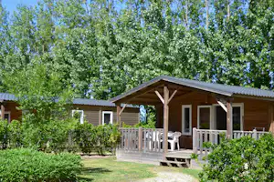 Holz-Ferienhaus mit Veranda, Sitzgelegenheit und Geländer, umgeben von Büschen und hohen Bäumen im Grünen.